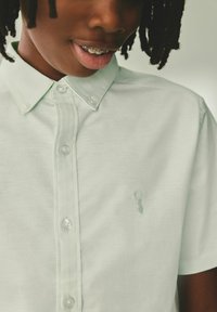Világoszöld pamut ing gombos gallérral, a bal mellrészen finom homárlogóval hímzett. Sima textúra és normál fazon.