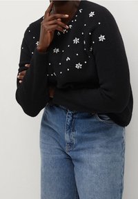 Pull noir avec des embellissements en forme d'étoile et de perles, tricot texturé, col rond, porté avec un jean en denim bleu clair.