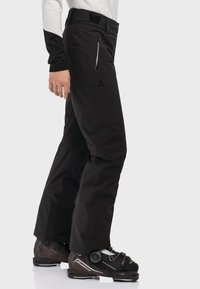 Schwarze Schneehose mit einem schlanken, geraden Schnitt. Verfügt über eine Reißverschlusstasche und ein dezentes Logo, kombiniert mit dunklen Skischuhen.