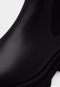 Mustast nahast pahkluu saabas sileda viimistlusega, elastsete külgpaneelide ja tugevast kummist tallaga. Omab ümarat varvast ja minimaalset õmblust.