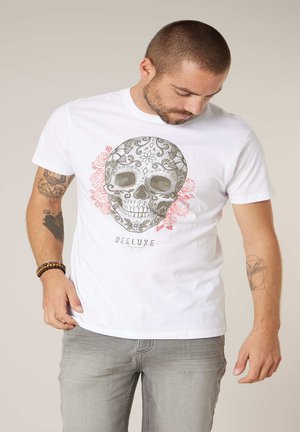 Homme aux cheveux courts et à la barbe portant un t-shirt blanc avec un imprimé de crâne décoré et de roses, un pantalon gris et des bracelets.