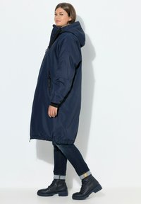 Abrigo impermeable azul marino con capucha, cuenta con un frente con cremallera, puños elásticos y dobladillo ajustable, combinado con jeans oscuros y botas negras.