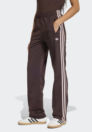 Persona vistiendo pantalones deportivos Adidas marrón oscuro con rayas rosa claro y zapatillas blancas con rayas negras, de pie con una mano en el bolsillo.