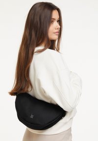 Borsa a tracolla nera, semi-circolare, con una texture liscia e un logo sottile. Indossata a spalla, abbinata a una felpa di colore chiaro.
