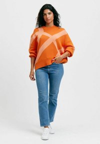 Orange tröja med rosa diagonala ränder, ballongsärmar och en rund halsringning, matchad med blå slim fit jeans och vita sneakers.