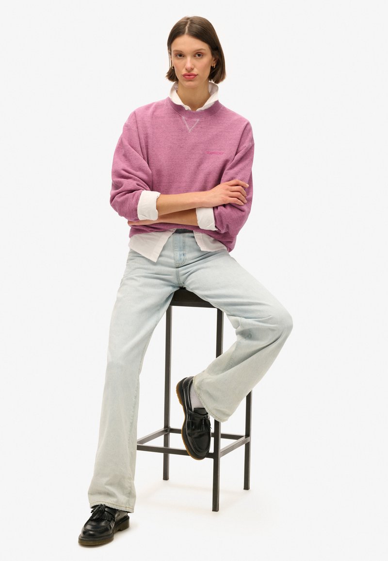 Maglione oversized viola chiaro con polsini a costine, indossato sopra una camicia bianca con colletto. Abbinato a jeans a gamba larga blu chiaro e scarpe nere.