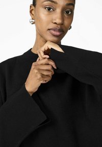 Maglione nero con scollo tondo e maniche larghe. Primo piano di mani che afferrano la manica, mostrando la texture a coste e i dettagli del tessuto.