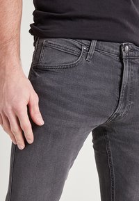 Hombre con jeans ajustados negros desgastados y una camisa negra, con la mano reposando a un lado.