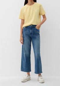 Camiseta de algodón amarilla con mangas cortas y un pequeño logo de corazón; combinada con jeans de mezclilla azul de pierna ancha y zapatillas blancas.