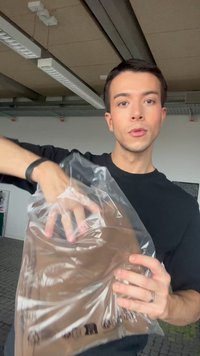 Νέος άνδρας με μαύρο πουκάμισο κρατάει και ανοίγει μια διάφανη πλαστική σακούλα που περιέχει ένα διπλωμένο καφέ ένδυμα σε εσωτερικό χώρο.