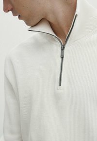 Pull blanc à fermeture éclair avec un design côtelé texturé. Dispose d'une fermeture à demi-zip et d'une fermeture éclair noire contrastante.