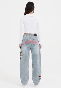 Hvid langærmet crop top og lyseblå baggy jeans med farverige broderede designs og rødt logo på ryggen. Blød denimtekstur.