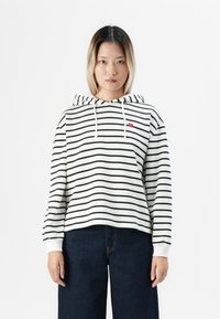 Jeune femme aux cheveux raides portant un sweat à capuche blanc avec des rayures noires et un petit cœur rouge, associé à un pantalon large en jean foncé.