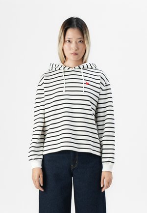 Jeune femme aux cheveux raides portant un sweat à capuche blanc avec des rayures noires et un petit cœur rouge, associé à un pantalon large en jean foncé.