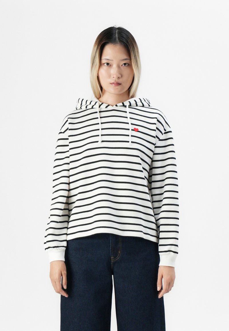 Jeune femme aux cheveux raides portant un sweat à capuche blanc avec des rayures noires et un petit cœur rouge, associé à un pantalon large en jean foncé.