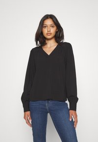 Blusa nera a scollo a V con maniche lunghe, dettagli plissettati sulle spalle, realizzata in tessuto leggero, abbinata a jeans blu.
