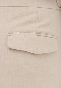 Beżowa tkanina o fakturowanej powierzchni z pojedynczą kieszenią naszywaną po prawej stronie. Szwy podkreślają kształt kieszeni.