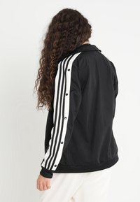 Persona con cabello largo y rizado, vistiendo una chaqueta de chándal negra con franjas blancas y botones a presión a lo largo de las mangas, vista desde atrás.