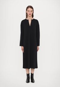 Black, knee-length dress with a V-neck and long sleeves, made from a smooth fabric. Paired with black, studded ankle boots.