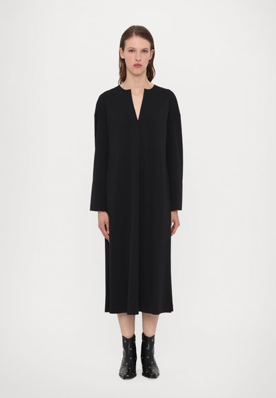Robe noire longueur genoux avec un décolleté en V et des manches longues, fabriquée à partir d'un tissu lisse. Associée à des bottines noires à clous.