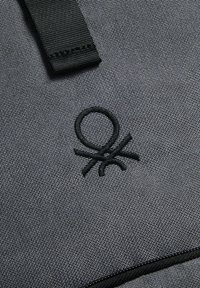 Logotipo bordado negro sobre un fondo de tela gris texturizada, con una correa negra resistente y costuras visibles.