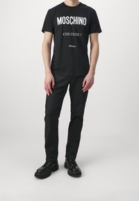 Man som bär en svart Moschino Couture t-shirt, svarta byxor och svarta läderskor med snörning, stående mot en enkel vit bakgrund.