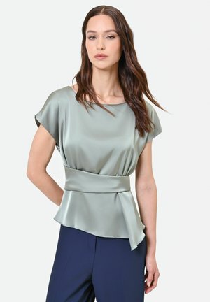 Donna che indossa una camicetta in raso verde chiaro con una fascia larga in vita e pantaloni blu navy, in piedi davanti a uno sfondo bianco uniforme.