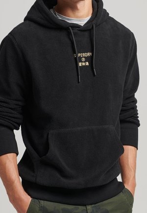 Homme portant un sweat à capuche en polaire noire avec un petit texte sur la poitrine et les mains dans les poches d’un pantalon camouflage.