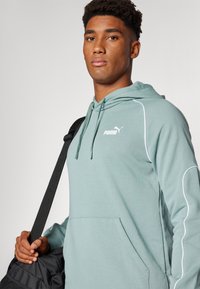 Svetlozelená mikina s bielym šitím a klokaním vreckom, s malým bielym logom Puma. Model drží čiernu tašku s popruhom.
