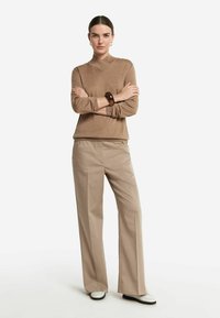 Beige tröja med korskänsla vid halsen, i kombination med ljusbeiga vida byxor. Noterbart iögonfallande träarmband på handleden.