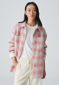 Vrouwelijke, rozige en grijze geruite shacket met twee voorzakken, knopen en een relaxed fit, gecombineerd met een witte blouse en crèmekleurige broek.