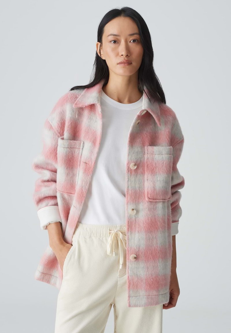 Vrouwelijke, rozige en grijze geruite shacket met twee voorzakken, knopen en een relaxed fit, gecombineerd met een witte blouse en crèmekleurige broek.