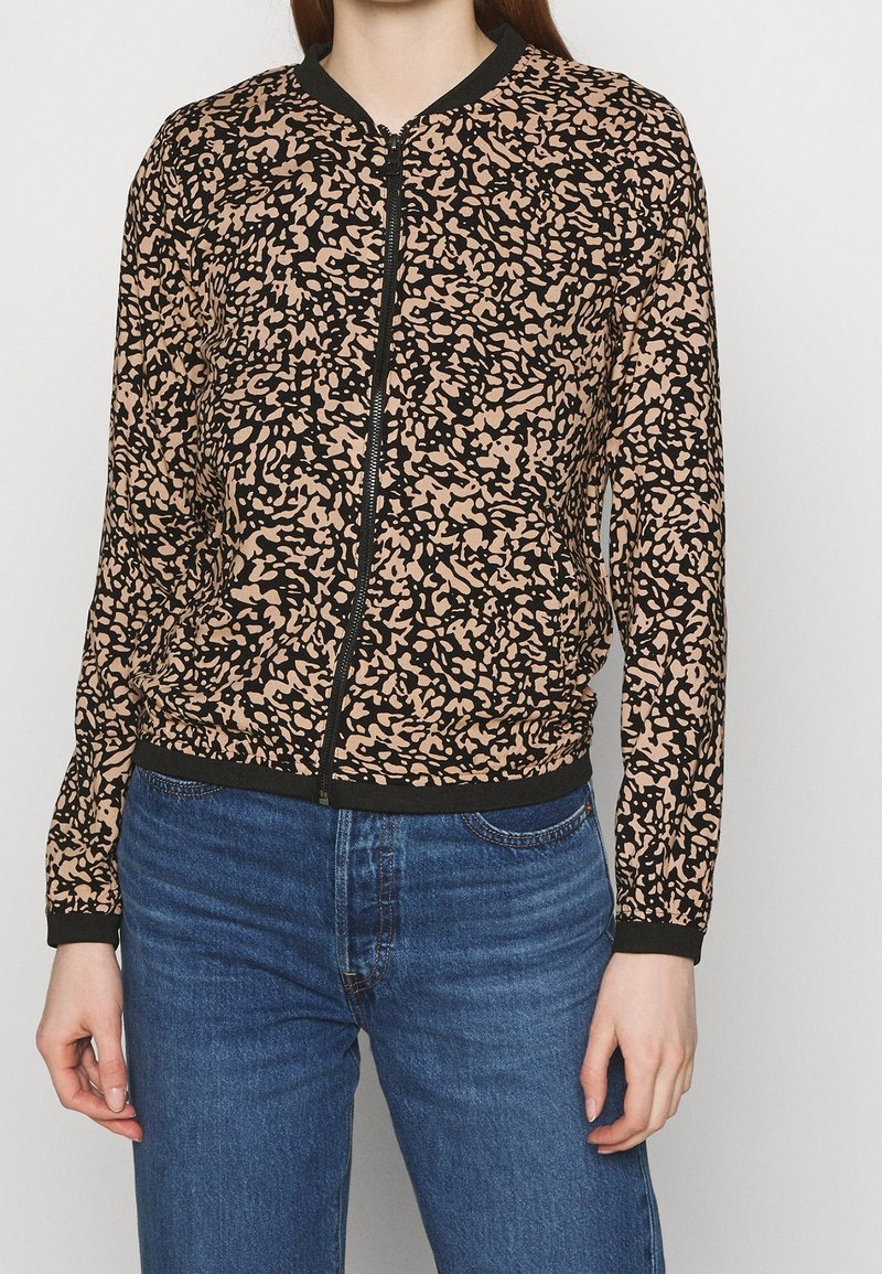 Femme portant une veste zippée à motif abstrait noir et beige avec des bordures noires et un jean en denim bleu, sur un fond uni.