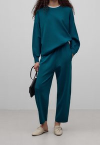 Turkusowy, oversize'owy sweter oraz dopasowane, zwężane spodnie, wykonane z gładkiego materiału, połączone z beżowymi klapkami z węzłem i małą czarną torebką.