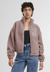 Osoba koja nosi mauve flis jaknu s džepom na prsima s patentom, crnu majicu i hlače visokog struka plave boje na jednobojnoj pozadini.