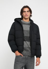 Giovane uomo con capelli castani mossi indossa un giubbotto imbottito nero sopra un maglione a righe grigie e nere e jeans grigi, in piedi con una mano in tasca.