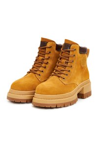 Suède gele enkelboots met bruine accenten, ronde neus, dikke rubberen zool en vetersluiting. Opvallend logo detail aan de zijkant.