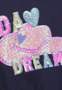 Kapelusz kowbojski pokryty cekinami, z sercami i wielokolorowymi literami układającymi się w słowo "Marzenie" na ciemnym tle.