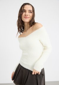 Weißer, schulterfreier Pullover aus flauschigem Material mit geripptem Kragen und Bündchen. Getragen mit einem dunklen, plissierten Rock mit sichtbarem Reißverschluss.