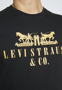 Camiseta de algodón negra con un diseño gráfico amarillo de dos caballos tirando de un par de pantalones, junto con el texto "LEVI STRAUSS & CO."