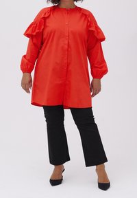 Camicia rossa con bottoni, maniche arricciate, vestibilità ampia e maniche lunghe. Abbinata a pantaloni neri a campana e tacchi neri a punta.