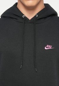 Juodas gobtuvėlis, pagamintas iš minkštos medžiagos, su kišenėle, primenančia kengūrą, raišteliais reguliuojamu gobtuvu ir mažu rožiniu Nike logotipu ant krūtinės.