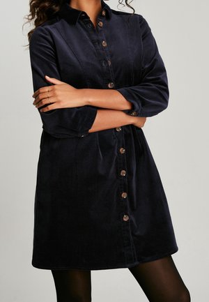 Marineblaues Corduroykleid mit knöpfbarem Vorderteil, langen Ärmeln, die bis zu den Ellenbogen gerollt sind, und einer lässigen Passform. Mit braunen Knöpfen und Taschen.