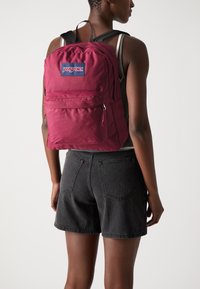 Sac à dos bordeaux avec des sangles noires ajustables, doté d'une poche zippée à l'avant et d'une étiquette logo rectangulaire. Texture de tissu lisse, design minimal.