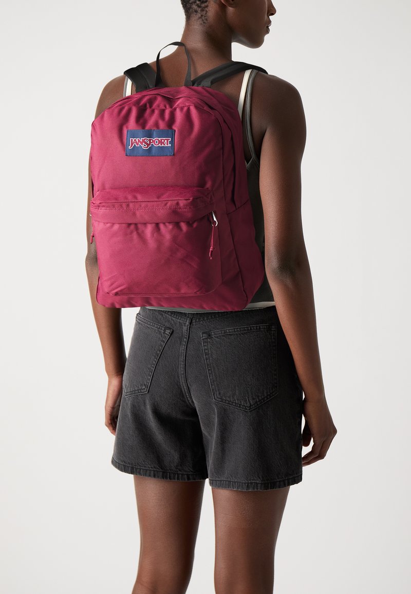 Sac à dos bordeaux avec des sangles noires ajustables, doté d'une poche zippée à l'avant et d'une étiquette logo rectangulaire. Texture de tissu lisse, design minimal.