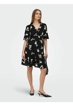 Kvinne iført en svart knelang kjole med hvitt blomstertrykk, svarte slingback hæler, holder en svart håndveske, står mot en ensfarget bakgrunn.