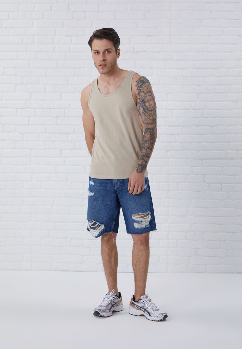 Beige tanktop, blå denimshorts med frynset kanter og flere revner, parret med sølv- og sorte træningssko. Stylisettet mod en hvid baggrund.