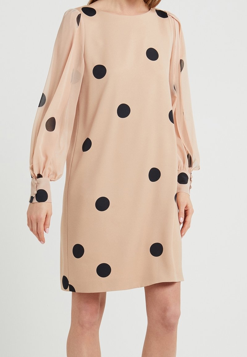 Robe beige à manches longues avec des pois noirs et des manches bouffantes transparentes, portée par une personne devant un fond clair uni.
