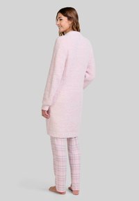 Mujer sonriendo, vistiendo un cárdigan largo y suave de color rosa claro y pantalones de pijama de cuadros rosas, de pie descalza frente a un fondo gris liso.