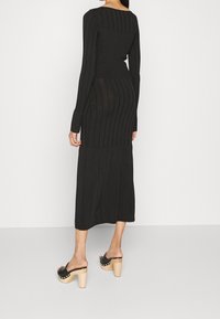 Donna che indossa un lungo abito nero a coste e scarpe con plateau nere con suole in legno, in piedi su uno sfondo bianco uniforme.